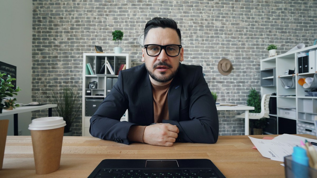 Team lead with glasses and a navy jacket seated at his desk during an office video call